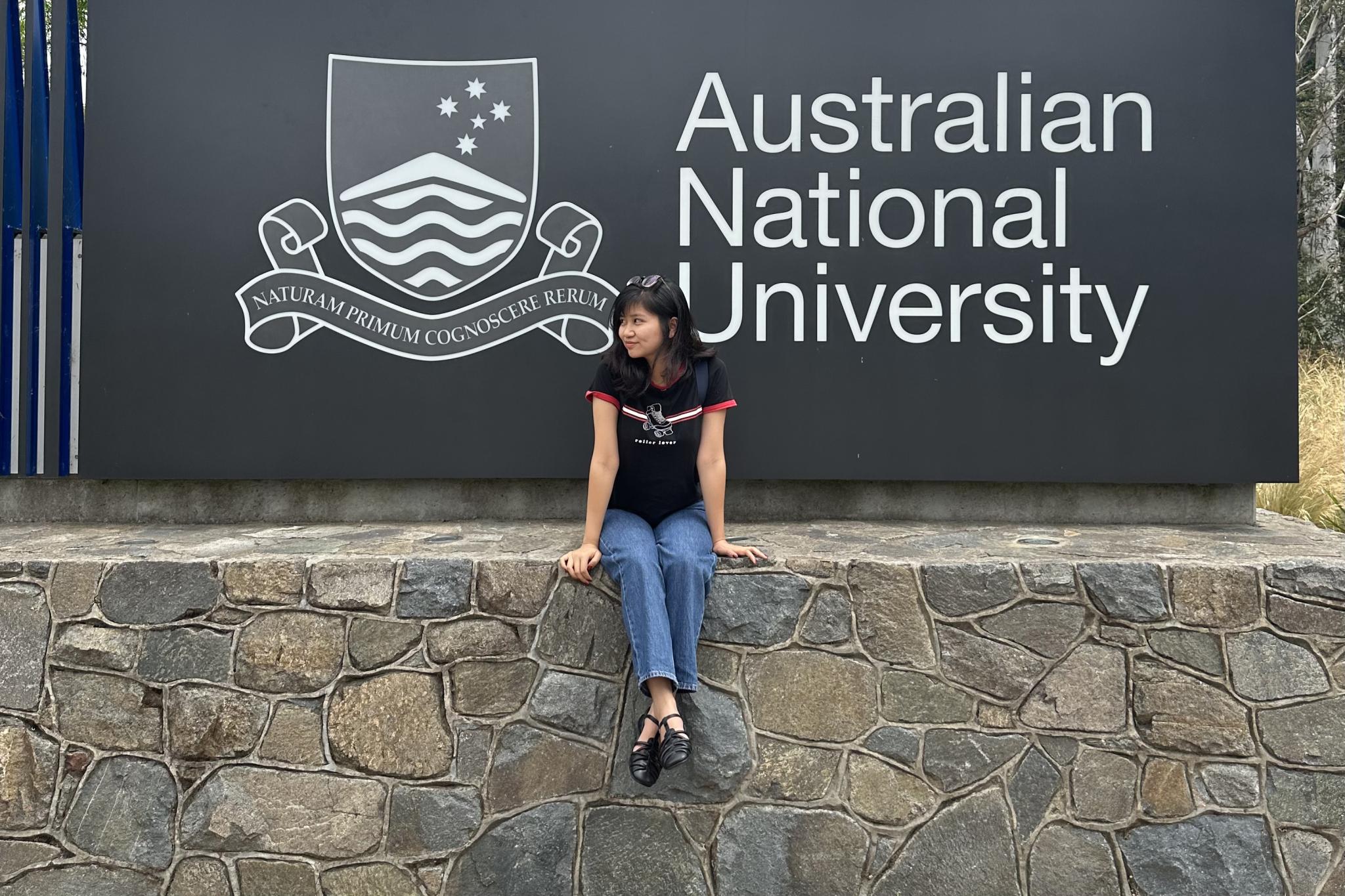 International postgraduate student Binh Thanh Ho sitting on a stone wall in front of a sign for the Australian National University